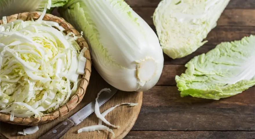 Frischer Chinakohl, bereit für leckere Chinakohl Rezepte. Ein ganzer Kohlkopf, geschnittene Blätter und fein gehackter Kohl in einem Korb liegen auf einem Holztisch.