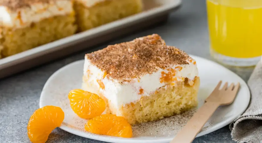 Ein saftiges Stück Fantakuchen-Rezept mit Mandarinen und Sahne, bestreut mit Zimt. Dazu ein Glas Fanta und frische Mandarinenstücke.