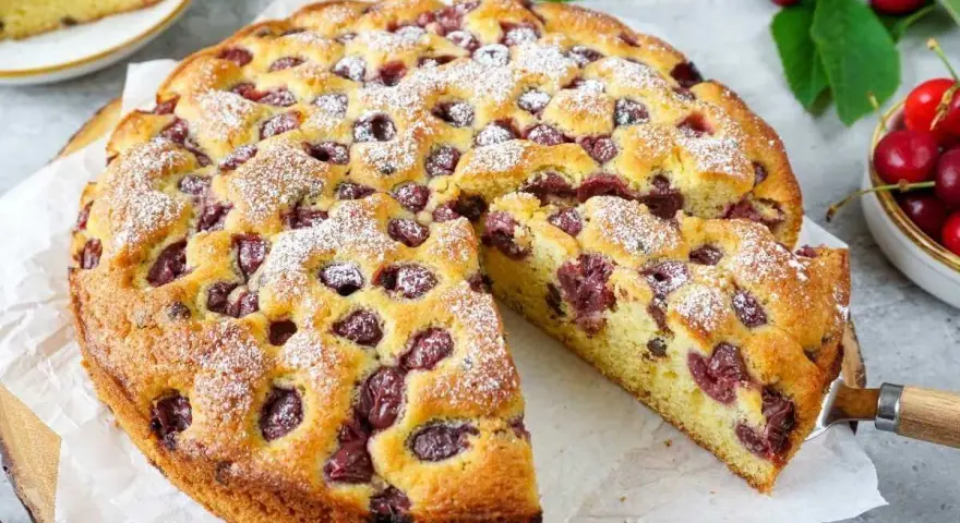 Ein saftiger Kirschkuchen, bestäubt mit Puderzucker, mit einem Stück herausgeschnitten. Frische Kirschen und Blätter liegen daneben.