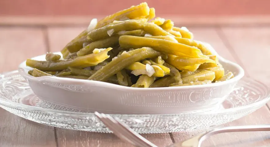 Omas Bohnensalat Rezept: Grüne Bohnen mit Zwiebeln in einer Schüssel auf einem Glasuntersetzer, daneben eine Gabel.