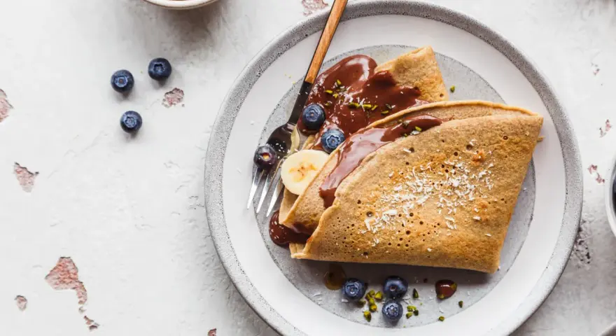 Buchweizen-Crêpes mit Schokoladensoße, Blaubeeren und Banane. Ein köstliches Beispiel für Buchweizen Rezepte, garniert mit Kokosraspeln und Pistazien.