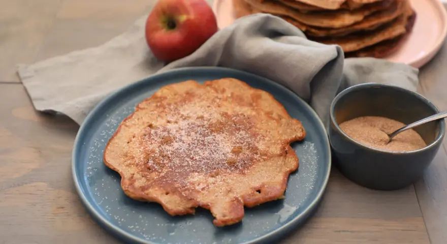 Ein Apfelpfannkuchen, bestreut mit Zucker und Zimt, liegt auf einem blauen Teller. Daneben ein roter Apfel und eine Schüssel mit Zimtzucker. Ein leckeres Apfelpfannkuchen Rezept zum Nachmachen.