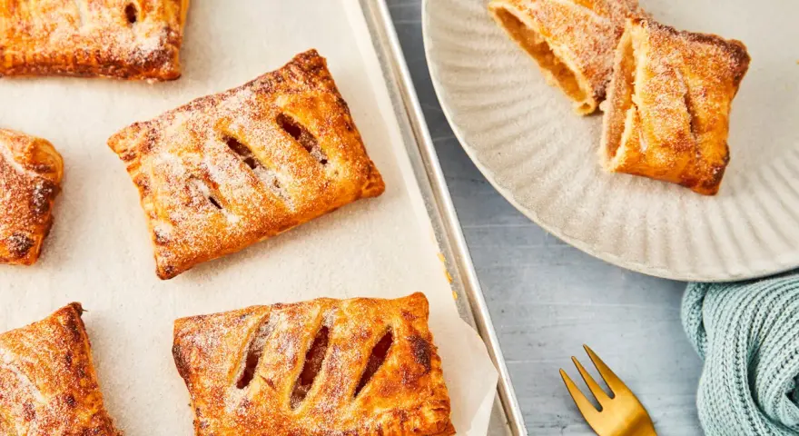 Goldbraune Apfeltaschen, bestäubt mit Puderzucker, warten auf dem Backblech und auf einem Teller. Ein köstliches Apfeltaschen Rezept zum Nachbacken.