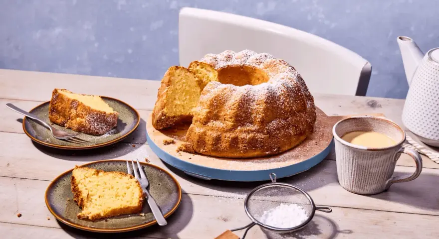 Ein goldbrauner Kartoffelkuchen mit Puderzucker bestreut, daneben zwei Teller mit Kuchenstücken, eine Tasse Kaffee und eine Kanne. Ein tolles Kartoffelkuchen Rezept!