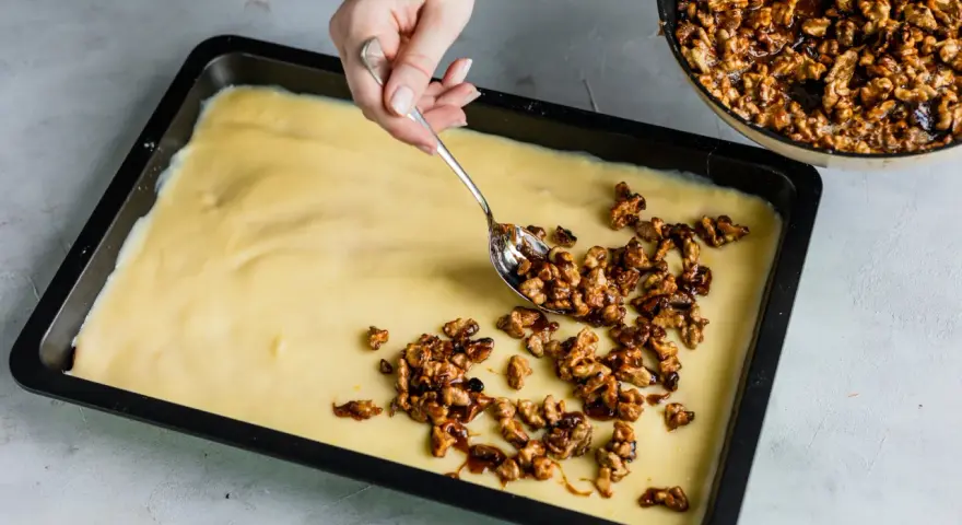 Eine Hand verteilt karamellisierte Walnüsse auf einem hellgelben Teig in einer Backform. Bald gibt es einen "friss dich dumm Kuchen"!