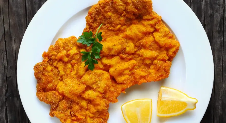 Ein knuspriges Schnitzel im Backofen, goldbraun paniert, mit Petersilie und Zitronenspalten auf einem weißen Teller serviert.