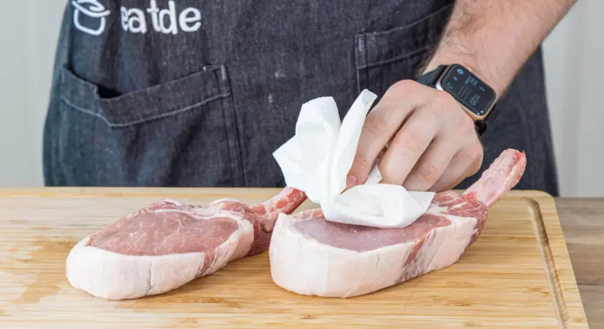 Vorbereitung für ein saftiges Kotelett Rezept: Frische Schweinekoteletts werden auf einem Holzbrett mit Küchenpapier abgetupft.