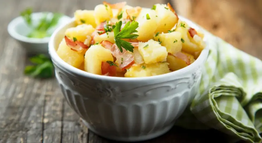 Ein einfacher Kartoffelsalat mit Speck und Petersilie in einer Schüssel, serviert auf einem Holztisch.