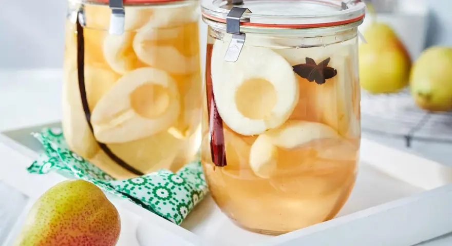 Zwei Gläser mit eingekochten Birnen, verfeinert mit Zimt und Sternanis. Ein frische Birne liegt daneben.