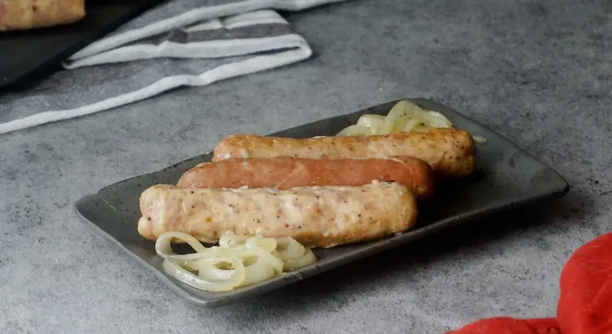 Drei Bratwürste mit Zwiebeln auf einem Teller, bereit zum Servieren. Sie sehen aus, als wären sie gerade aus dem Backofen gekommen.