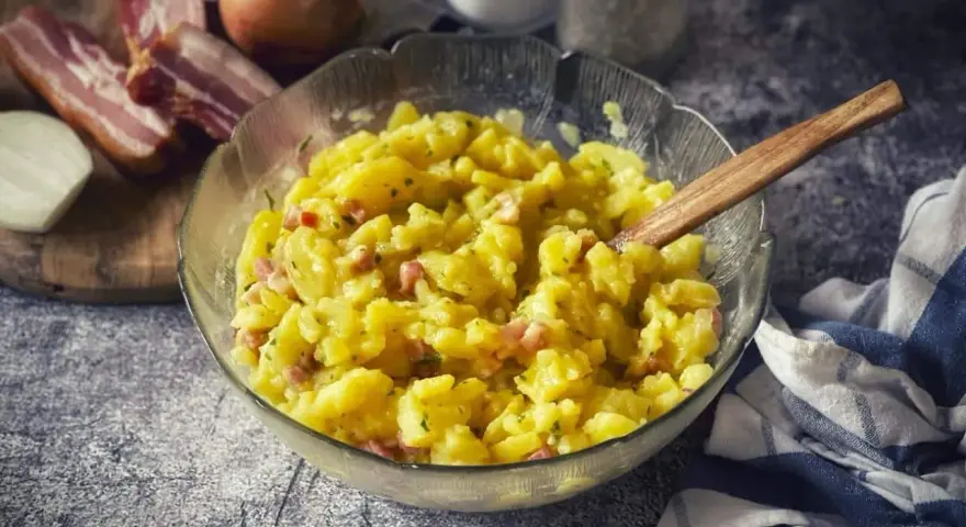 Schüssel mit warmem Kartoffelsalat, garniert mit Speck und Kräutern, daneben Zwiebeln und Speck.