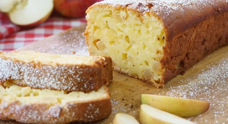 Ein saftiger, schneller Apfelkuchen mit Puderzucker bestäubt, daneben Apfelspalten und ganze Äpfel auf kariertem Tuch.