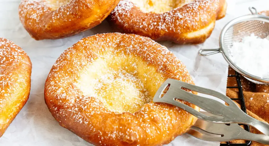 Frisch gebackene, ausgezogene Krapfen, bestäubt mit Puderzucker. Ein Kochlöffel hält einen der goldbraunen Krapfen.