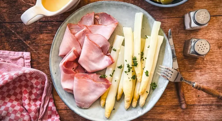 Spargel mit Schinken, garniert mit Petersilie, auf einem Teller mit Sauce Hollandaise und Kartoffeln daneben.