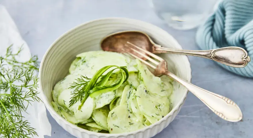 Omas Gurkensalat mit Joghurt, cremig und frisch, garniert mit Dill. Ein Löffel und eine Gabel liegen bereit.