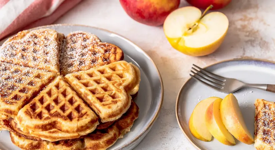 Herzförmige Waffeln mit Puderzucker, daneben frische Äpfel und Apfelspalten. Ein Genuss für alle Sinne!