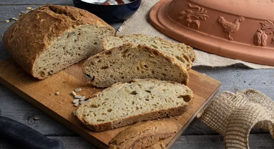 Frisch gebackenes Dinkelbrot mit Körnern, aufgeschnitten auf einem Holzbrett. Daneben ein Römertopf, eine Schüssel mit Joghurt und ein Messer.