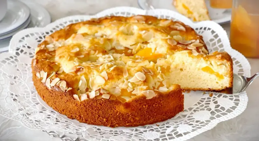 Ein goldbrauner Landfrauen Aprikosenkuchen, bestreut mit Mandelblättchen, wird auf einer zarten Tortenplatte serviert. Ein Stück wird gerade herausgehoben.