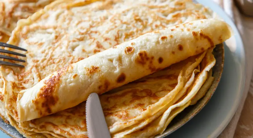 Ein Stapel goldbrauner Pfannkuchen, einer davon aufgerollt, mit Messer und Gabel. Perfekt für ein leckeres Pfannkuchen Rezept!