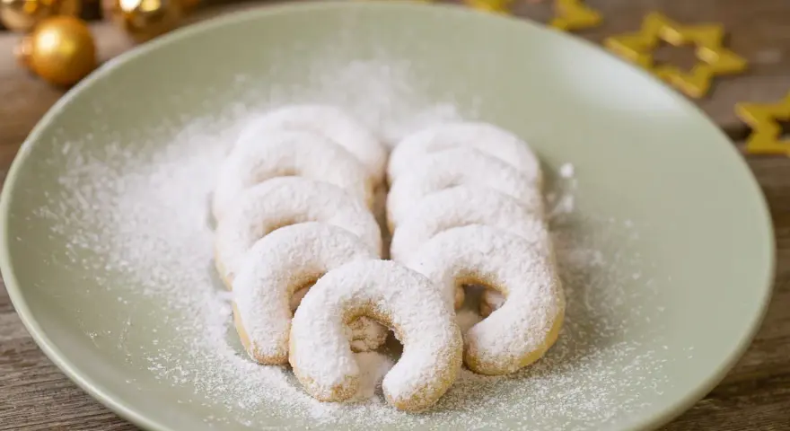 Vanillekipferl, bestäubt mit Puderzucker, liegen auf einem Teller. Goldene Kugeln und Sterne im Hintergrund deuten auf Weihnachten hin.