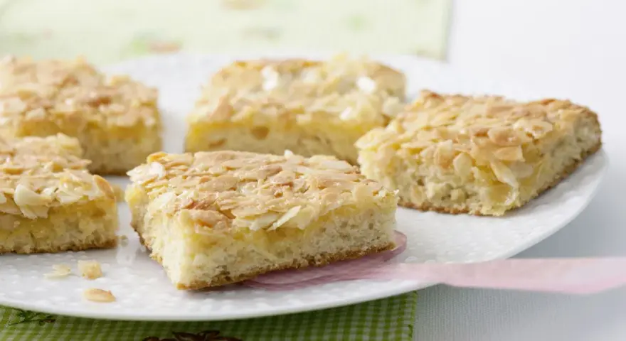 Saftiger mandelkuchen vom blech, goldbraun gebacken und mit knusprigen Mandelblättchen bestreut, liegt auf einer weißen Platte.
