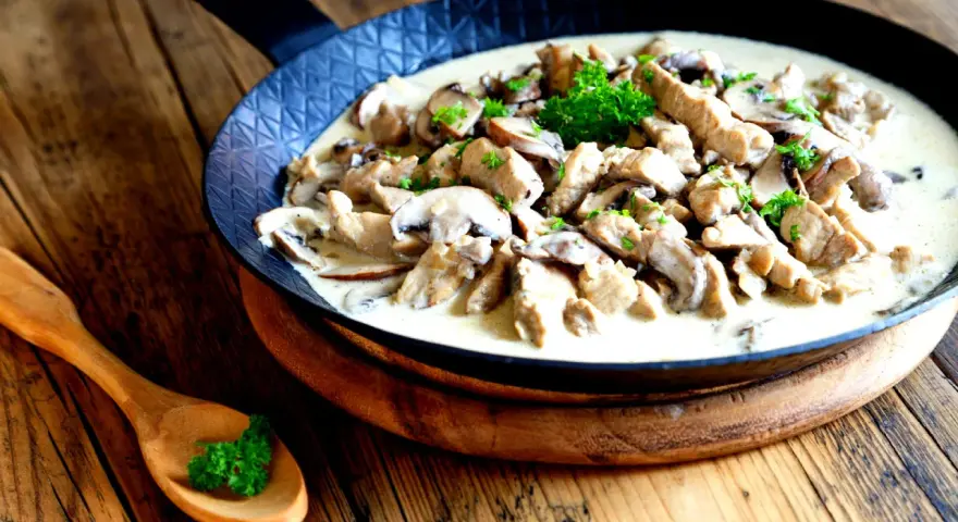 Putengeschnetzeltes mit Pilzen in cremiger Soße, garniert mit Petersilie, serviert in einer Pfanne auf rustikalem Holztisch.