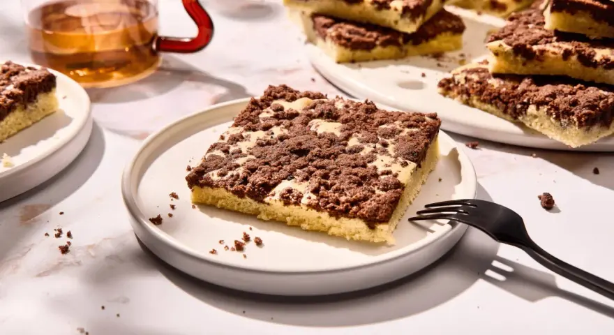 Ein Stück versunkener Streuselkuchen mit Schokostreuseln auf einem Teller, daneben eine Gabel und ein Glas Tee.