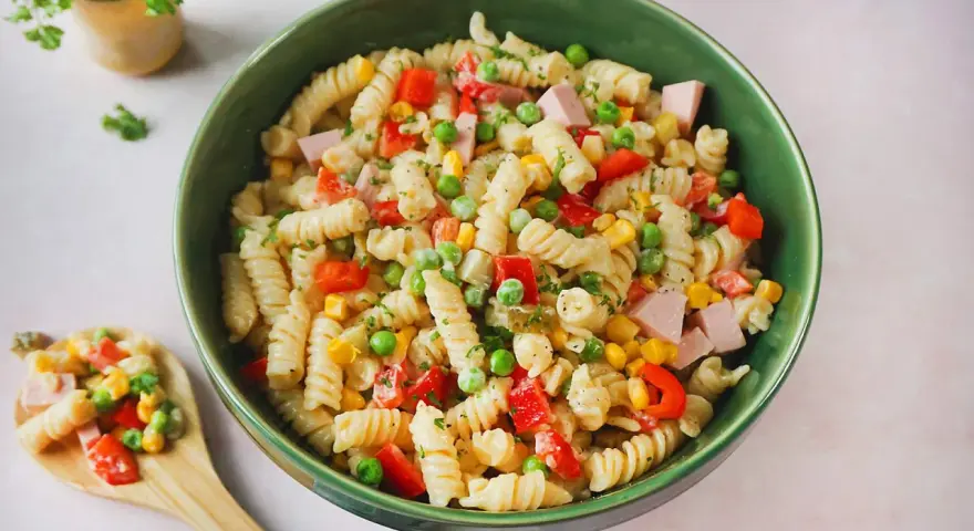 Ein bunter Nudelsalat mit Fleischwurst, Erbsen, Mais und Paprika in einer grünen Schüssel.
