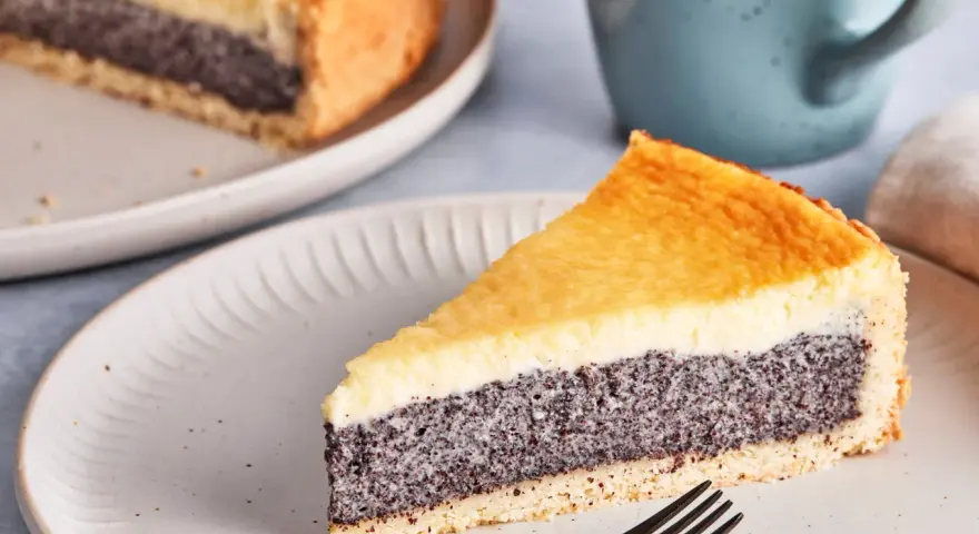 Ein Stück Mohn Schmand Kuchen mit goldbraunem Boden und cremiger Füllung auf einem Teller, daneben eine Tasse.