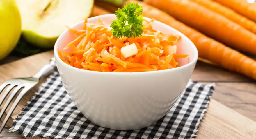 Frischer Karotten-Apfel-Salat in einer weißen Schüssel, garniert mit Petersilie. Im Hintergrund liegen ganze Karotten und ein halber Apfel.