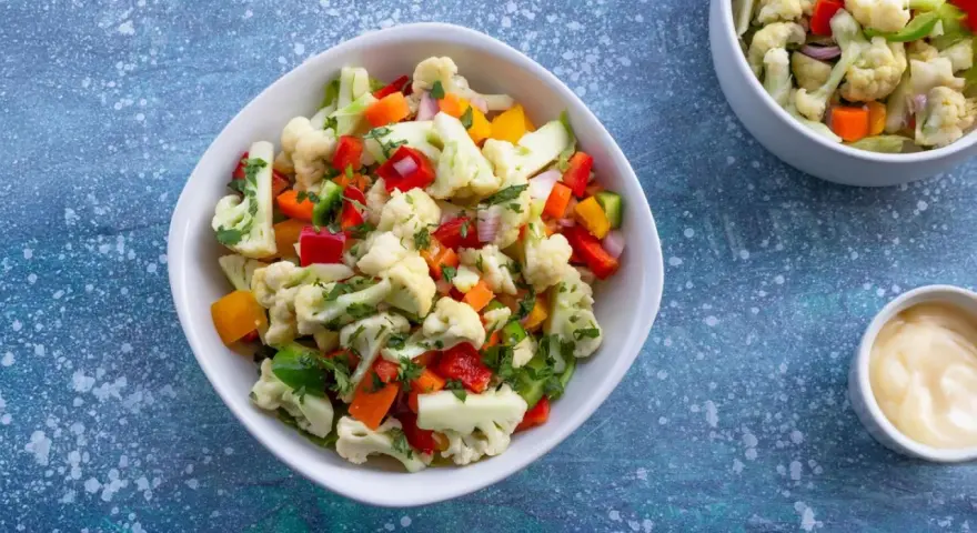Frischer blumenkohl salat mit Paprika, Karotten und Kräutern, serviert in einer weißen Schüssel. Daneben eine kleine Schale mit Dip.