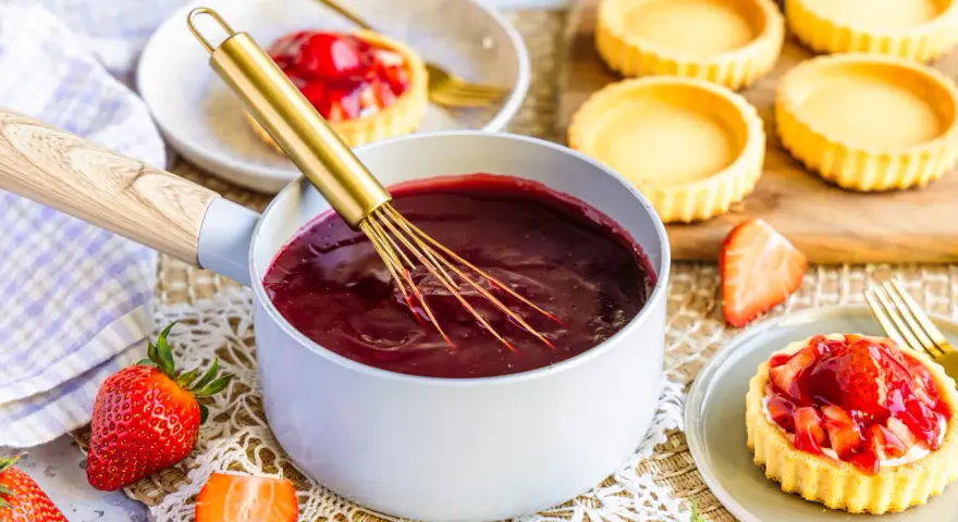 Frisch zubereiteter Tortenguss in einem Topf, bereit, um Obstkuchen zu verfeinern. Daneben liegen frische Erdbeeren und leere Tartelettes.