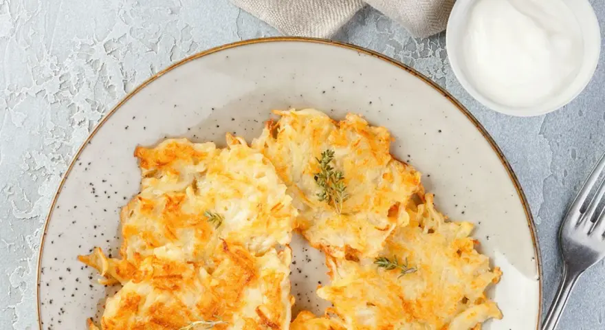 Knusprige Kartoffelpuffer, im Waffeleisen goldbraun gebacken, liegen auf einem Teller. Dazu ein Schälchen mit Sour Cream und ein Gabel.