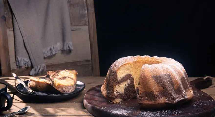 Ein fluffiger Marmorkuchen, bestäubt mit Puderzucker, liegt auf einem Holzbrett. Daneben zwei Stücke auf einem Teller.