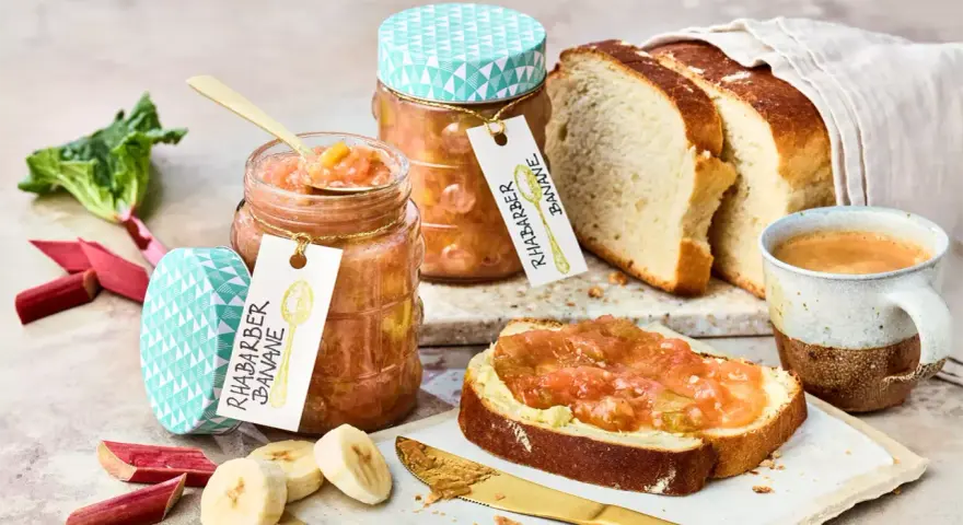 Zwei Gläser Rhabarber Bananen Marmelade, frischer Rhabarber, Bananenscheiben, Brot und Kaffee.