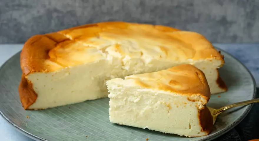 Ein saftiger Quarkkuchen ohne Boden, goldbraun gebacken, liegt auf einem Teller. Ein Stück ist bereits angeschnitten.