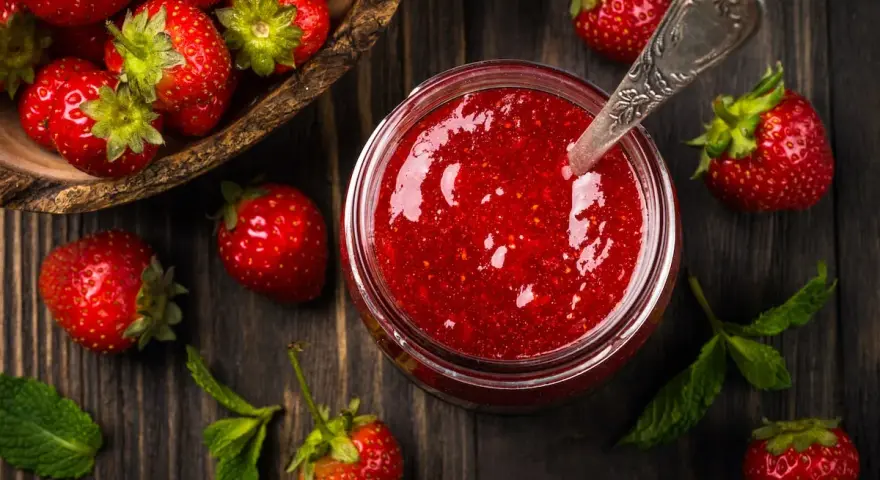 Frische Erdbeeren und ein Glas selbstgemachte Erdbeermarmelade, bereit zum Einkochen. Ein Löffel steckt in der Marmelade.