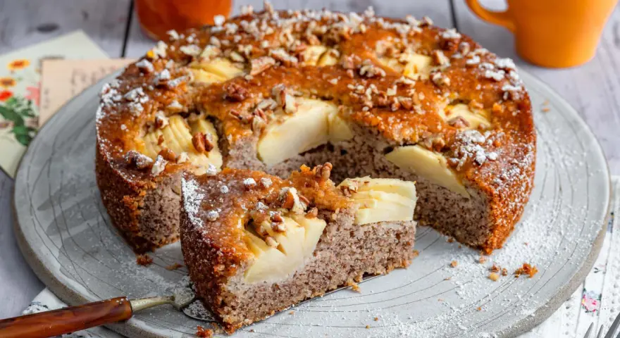 Ein saftiger Apfel-Birnen-Kuchen, bestreut mit Nüssen und Puderzucker, wird auf einem Teller serviert. Ein Stück ist bereits herausgeschnitten.