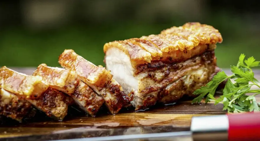 Knuspriger Schweinebauch, perfekt für ein köstliches schweinebauch rezept. Saftig, mit krosser Schwarte, auf einem Holzbrett mit Petersilie und einem Messer.