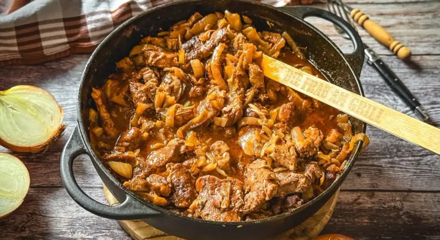 Herzhaftes Zwiebelfleisch-Rezept in Gusseisenpfanne, mit Zwiebeln und Fleischstücken in einer würzigen Sauce. Ein Holzlöffel mit der Aufschrift "Die Frau am Grill" liegt darin.