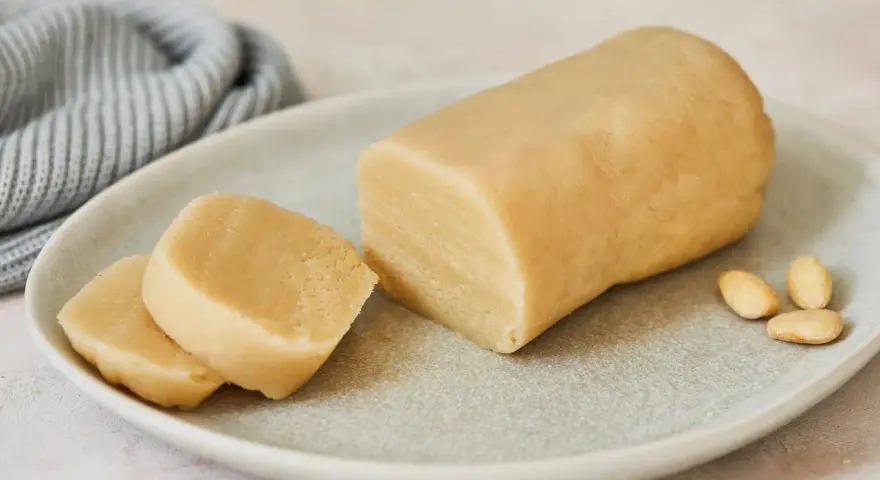 Frisch zubereitetes Marzipan zum **Marzipan selbst machen**. Ein Block und zwei Scheiben liegen auf einem Teller, daneben Mandeln.