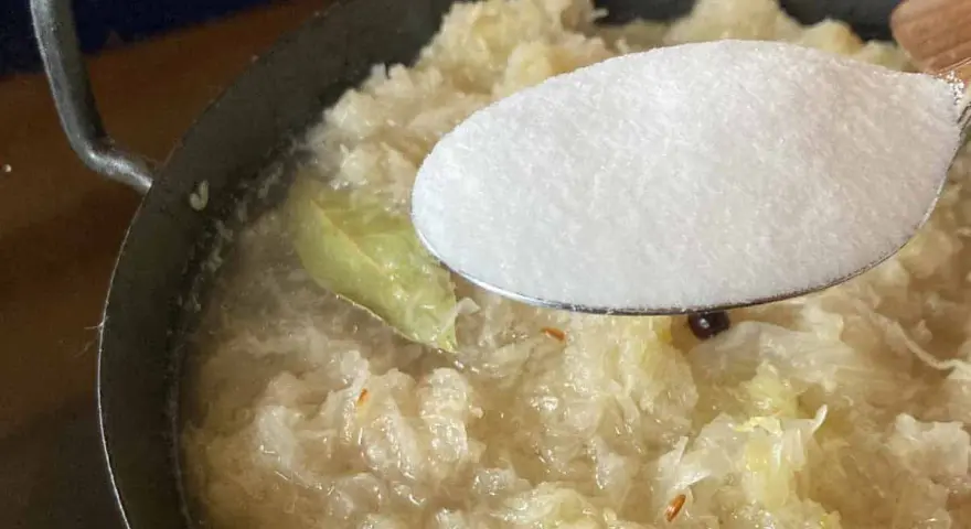 Zucker wird zu köchelndem Sauerkraut nach Omas Rezept gegeben. Ein Lorbeerblatt schwimmt im Topf.
