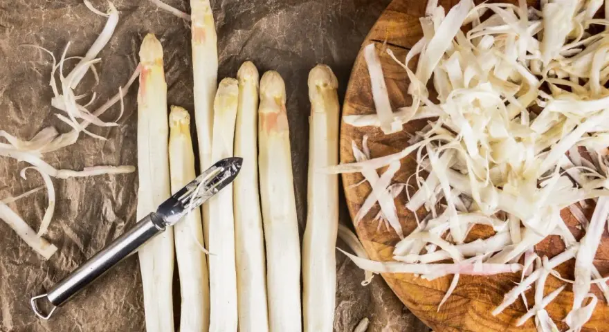 Frischer weißer Spargel wird geschält, die Spargelschalen liegen daneben. Ein Sparschäler liegt auf den Spargelstangen.