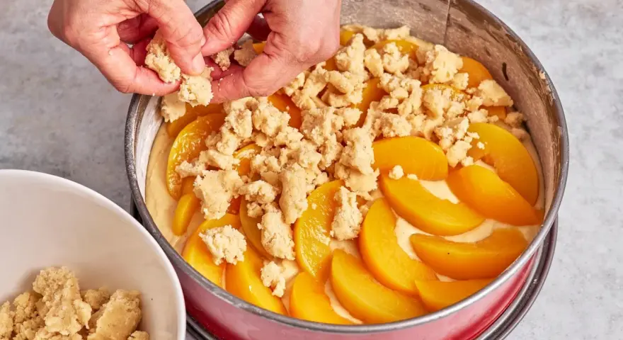 Hände verteilen Streusel auf einem Pfirsichkuchen in einer Springform.