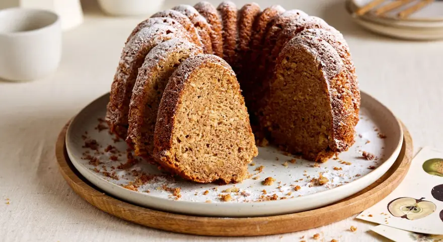 Ein saftiger Kuchen mit Dinkelmehl, bestäubt mit Puderzucker, liegt auf einem Teller. Ein Stück ist herausgeschnitten.