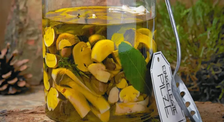 Glas mit eingelegten Pilzen, Kräutern und Lorbeerblatt. Ein Etikett mit der Aufschrift "Eingelegte Pilze" hängt am Glas.