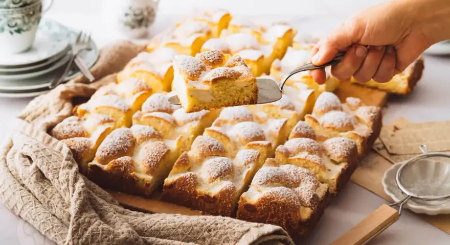 Ein Stück Spiegeleikuchen mit Quark wird aus einem Blechkuchen mit Puderzucker gehoben.
