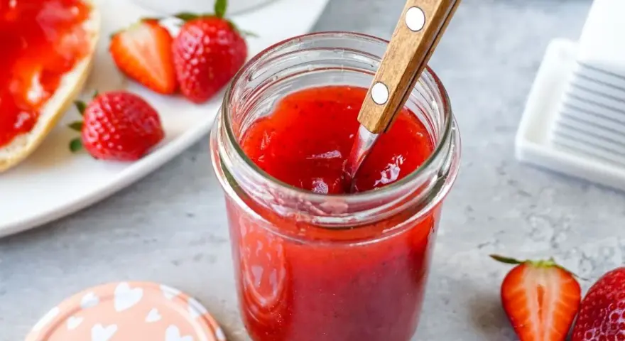 Ein Glas mit leuchtend roter Erdbeermarmelade, die mit einem Holzlöffel umgerührt wird. Daneben frische Erdbeeren und ein Stück Brot mit Marmelade – ein Genuss mit Pfiff!