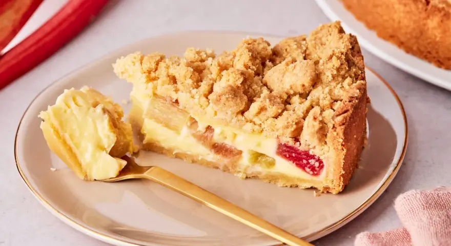 Ein Stück von Omas Rhabarberkuchen mit Pudding und Streuseln auf einem Teller, daneben ein goldener Löffel und rote Rhabarberstangen.