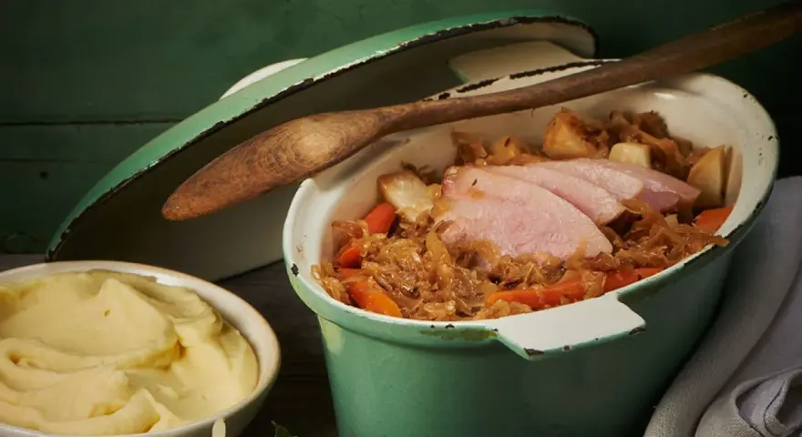 Herzhafter Szegediner Gulasch mit zarten Fleischscheiben, Sauerkraut und Karotten in einem grünen Topf, daneben eine Schüssel cremiges Kartoffelpüree.